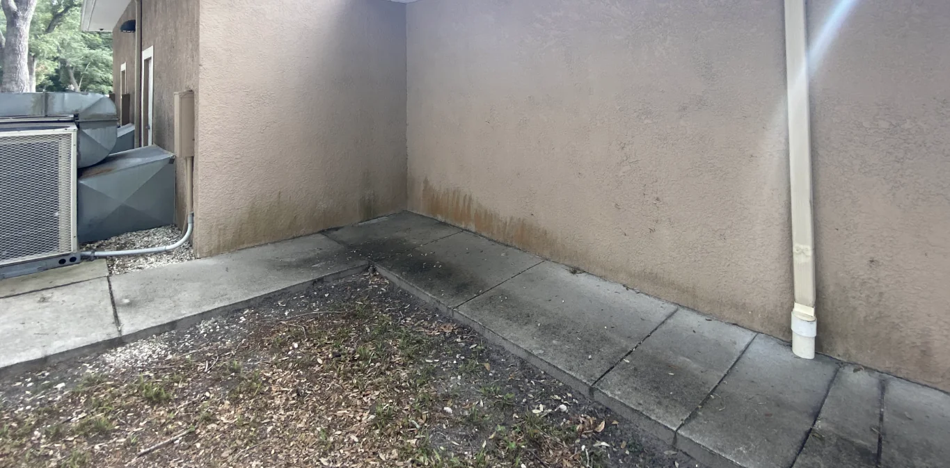 Stucco wall corner with dark and rust-colored staining below a downspout and mechanical area